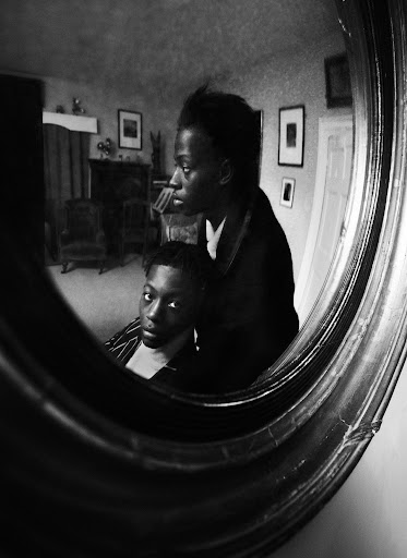 Black and white portrait of an African American mother and child reflected in an ornate mirror, set in a vintage-style home interior.