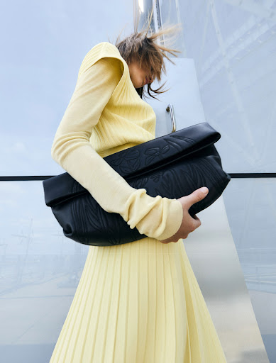 Fashion shoot featuring a female model in a pleated yellow skirt and long-sleeved top, holding a black leather embossed handbag outdoors.