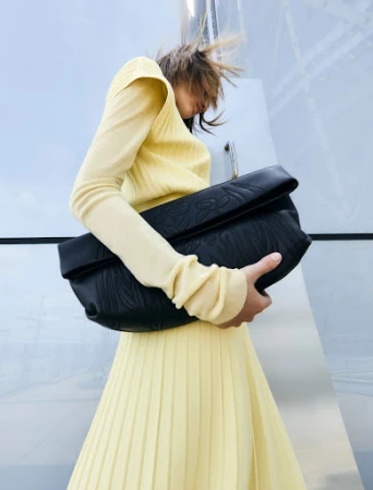 Fashion shoot featuring a female model in a pleated yellow skirt and long-sleeved top, holding a black leather embossed handbag outdoors.