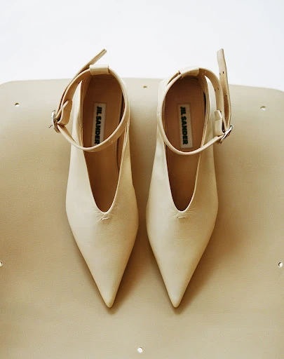 An overhead studio shot showcases a pair of light beige leather Jil Sander heeled shoes with pointed toes and ankle straps on a light background.
