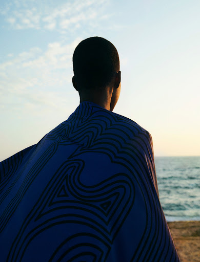 Adult with short hair wearing patterned shawl overlooking the ocean under a blue sky at sunset.