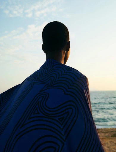 Adult with short hair wearing patterned shawl overlooking the ocean under a blue sky at sunset.