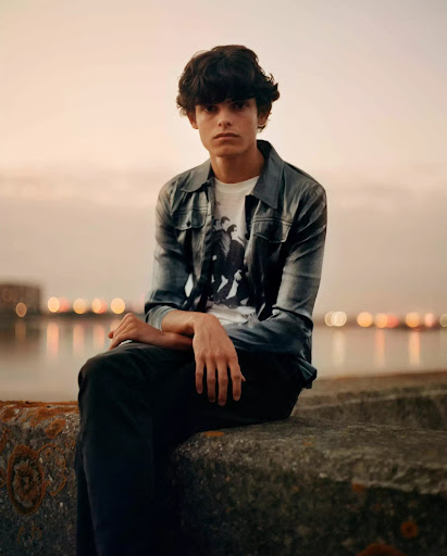 Young man with dark hair sitting on a stone wall near a body of water with city lights in the background, wearing casual clothing.