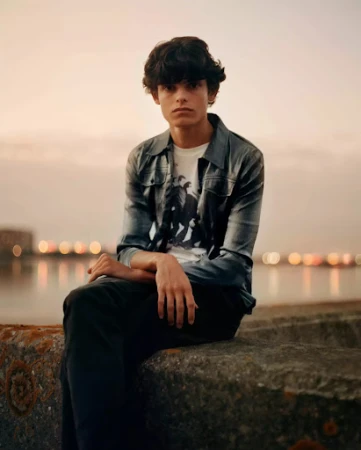 Young man with dark hair sitting on a stone wall near a body of water with city lights in the background, wearing casual clothing.