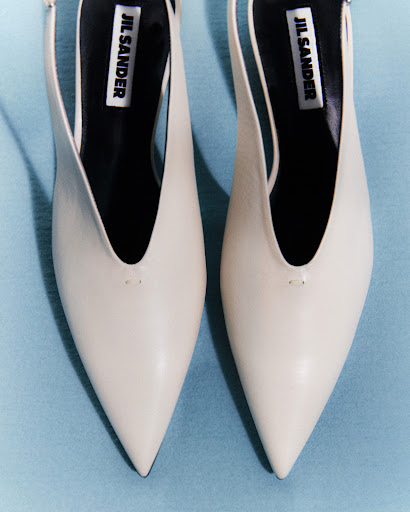 Close-up studio shot of a pair of Jil Sander slingback heels in off-white on a blue background, emphasizing their pointed toe and design.