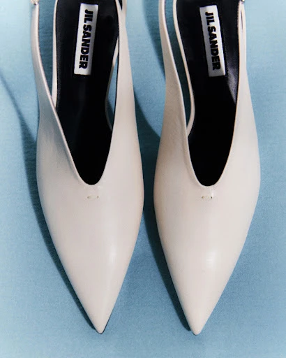 Close-up studio shot of a pair of Jil Sander slingback heels in off-white on a blue background, emphasizing their pointed toe and design.