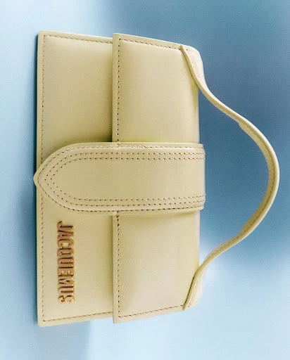 Close-up of a light yellow leather Jacquemus handbag with visible gold lettering on a light blue backdrop. Fashion accessory.