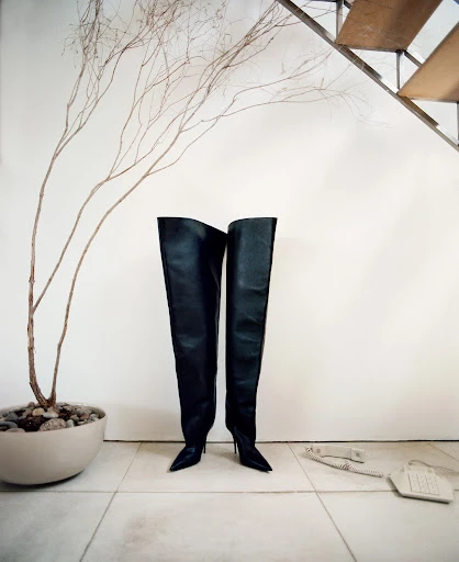 Pair of black, over-the-knee stiletto boots are displayed on a marble floor, set against a minimalistic background. Dried floral arrangement is visible.