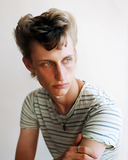 Portrait of a young adult male with blue eyes and distinctive blonde and dark hair styled in a pompadour wearing a striped shirt.