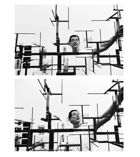 Black and white portrait of a man standing amidst a geometric metal sculpture. The man is wearing a white t-shirt in an indoor setting.