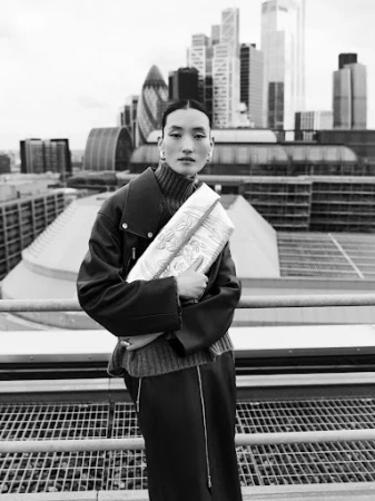 Monochrome outdoor fashion shot of a woman holding a decorative clutch bag, with a cityscape backdrop on an overcast day.