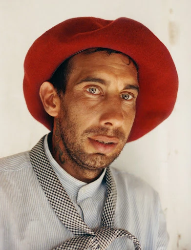 Close-up portrait of a man wearing a red beret, light blue striped shirt, and gingham tie, set against a textured white wall background.