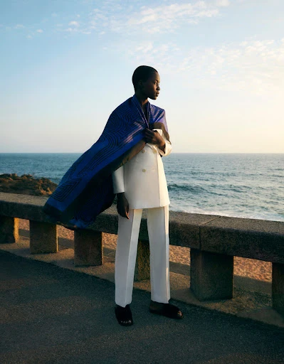 Fashion shot of a black woman in a white suit with a blue wrap standing on a stone wall near a beach with the ocean and sky in the background.