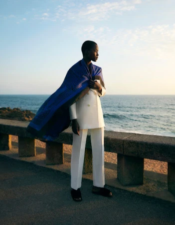 Fashion shot of a black woman in a white suit with a blue wrap standing on a stone wall near a beach with the ocean and sky in the background.