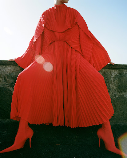 Fashion shot of a slender woman wearing a vibrant red pleated dress with long sleeves and matching high heel boots against a blue sky.