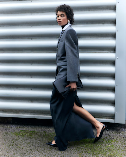 A woman strides confidently in a grey blazer, long black skirt with a slit, and pointy heels against a corrugated metal wall.