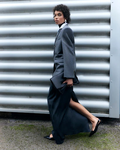 A woman strides confidently in a grey blazer, long black skirt with a slit, and pointy heels against a corrugated metal wall.