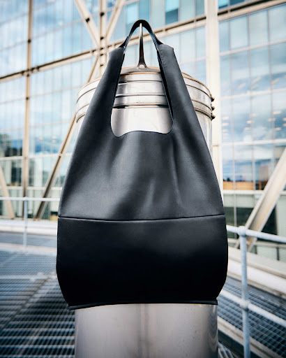 Black tote bag hangs from a silver metal post, set against a modern building with a windowed facade in an urban setting.
