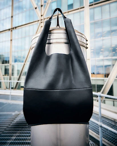 Black tote bag hangs from a silver metal post, set against a modern building with a windowed facade in an urban setting.