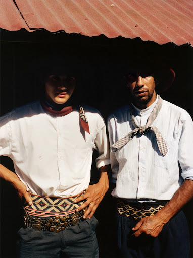 Two gauchos in traditional attire stand under a corrugated metal roof. One wears a red scarf, the other a patterned necktie.