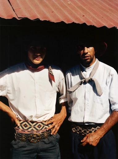 Two gauchos in traditional attire stand under a corrugated metal roof. One wears a red scarf, the other a patterned necktie.