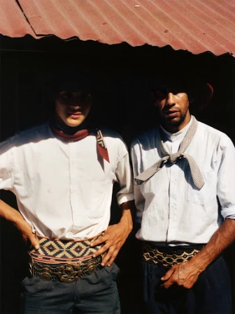 Two gauchos in traditional attire stand under a corrugated metal roof. One wears a red scarf, the other a patterned necktie.