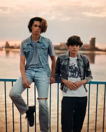 Two young men leaning on a blue metal railing outdoors against a sunset skyline with water.