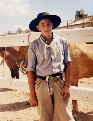 Portrait of a young adult male in traditional gaucho attire with a horse in the background, standing outside on a sunny day.