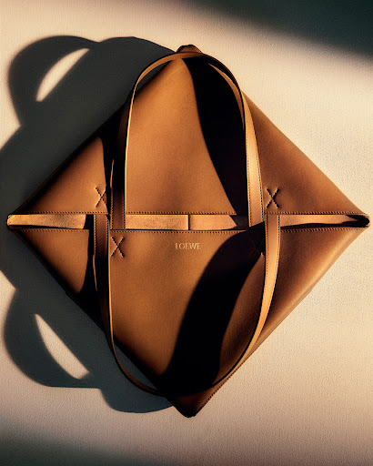 Close-up studio shot of a brown leather tote bag with top handles, casting a shadow on a light beige background. Loewe logo is visible.