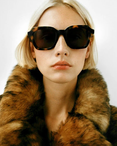 A woman with blonde hair wears tortoiseshell sunglasses and a large fur coat against a plain white background in a studio portrait.