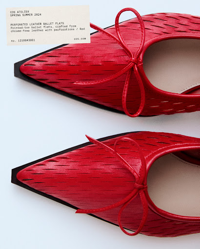 Close-up studio shot of two red pointed-toe ballet flats with bows, displayed on a white surface with a label visible in the background.