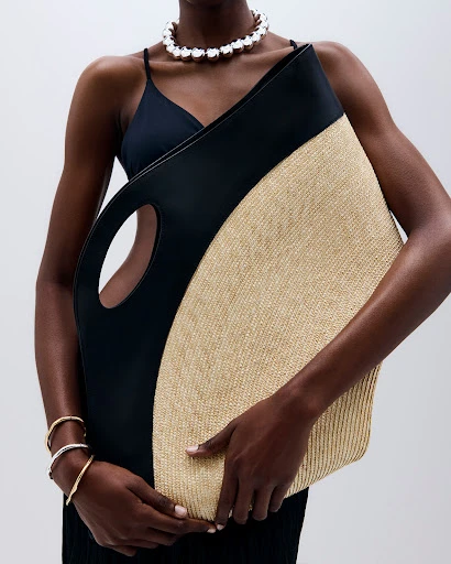 Woman in black dress holds black and beige straw tote bag and wears a pearl necklace and bracelets in a studio setting.