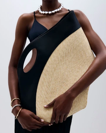 Woman in black dress holds black and beige straw tote bag and wears a pearl necklace and bracelets in a studio setting.