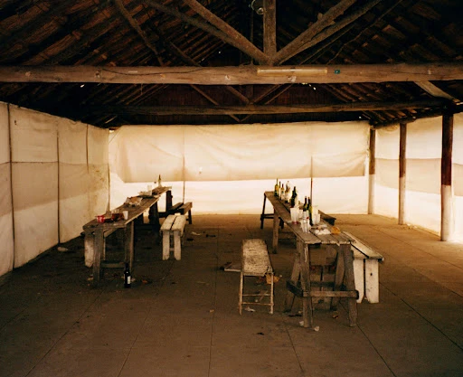 Rustic indoor space featuring wooden tables and benches, likely a dining area, with wine bottles and simple decor, under a wooden roof.