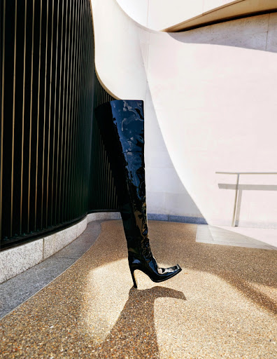 Black shiny thigh-high stiletto boot with a square toe sits on a speckled surface, against a white and dark wall.
