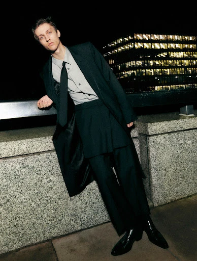 Man in black suit poses at night in front of an office building. He is leaning against a grey wall with black shoes and a black tie.