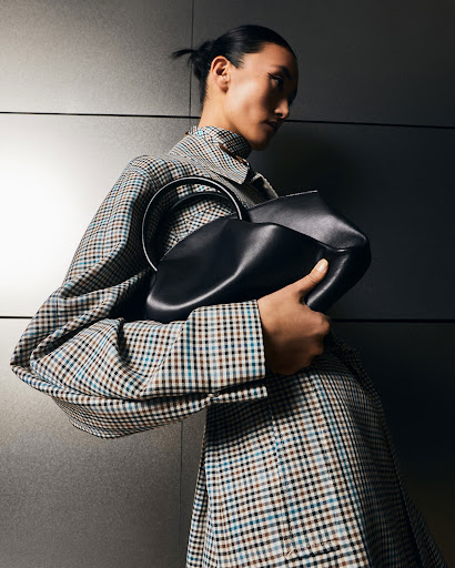 A woman in a checked coat holds a black leather handbag with a circular handle, posing against a neutral wall.