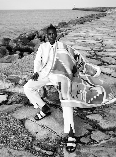 Black and white portrait of a Black male in an all-white suit sitting on a stone pier near the ocean, wearing sandals and holding a patterned textile.