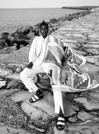 Black and white portrait of a Black male in an all-white suit sitting on a stone pier near the ocean, wearing sandals and holding a patterned textile.