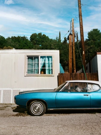 Aqua classic car parked outside a mobile home with a child looking out the window, utility poles, trees, and blue cloudy sky.