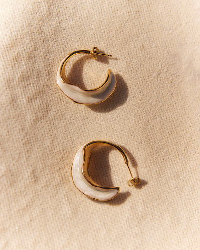 A pair of gold hoop earrings with white enamel accents are displayed on a textured beige fabric in soft, natural lighting.