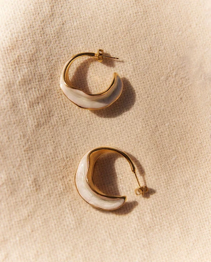 A pair of gold hoop earrings with white enamel accents are displayed on a textured beige fabric in soft, natural lighting.