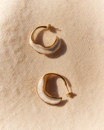 A pair of gold hoop earrings with white enamel accents are displayed on a textured beige fabric in soft, natural lighting.