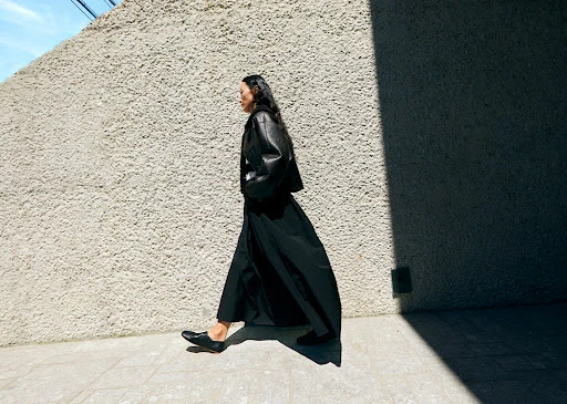 Model in black leather jacket and long black skirt walking past a textured concrete wall outdoors.