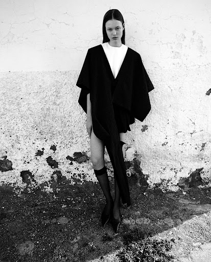 Monochrome fashion portrait of a model wearing a black cape over a white top and sheer black stockings against a textured wall.