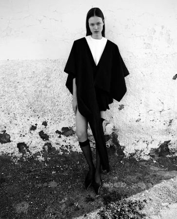 Monochrome fashion portrait of a model wearing a black cape over a white top and sheer black stockings against a textured wall.