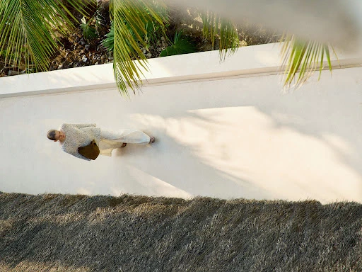 Eye-level aerial view of a woman in white loungewear walking along a white wall next to the green palm fronds and a thatched roof.