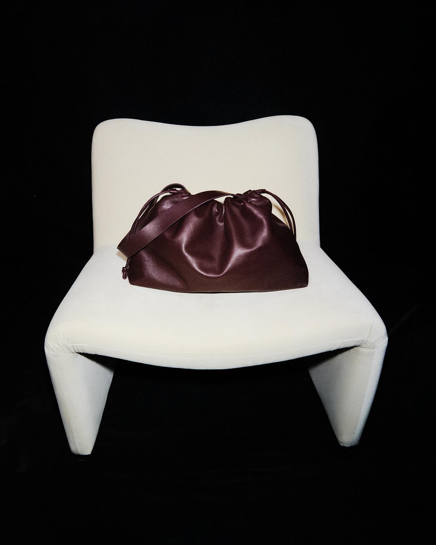Auburn leather drawstring bag sits on a modern white chair against a black background in a fashion product-style photograph.