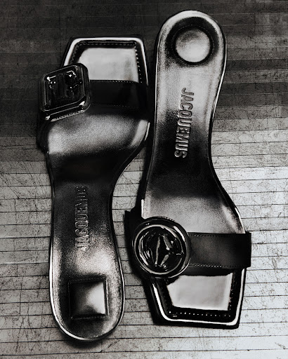 Shiny Jacquemus women's sandals in black and white, presented at an upward angle on a wood-grain surface. Stylish, modern footwear.