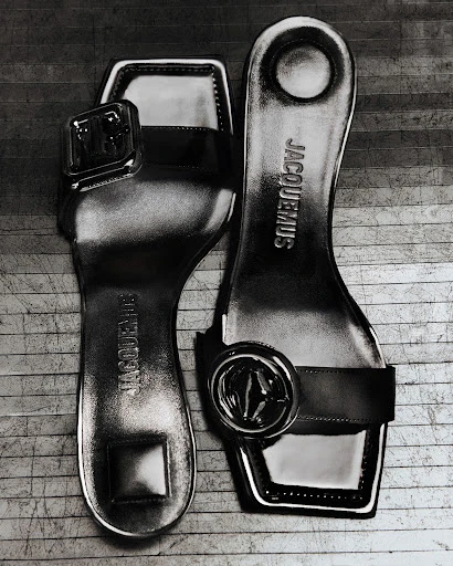 Shiny Jacquemus women's sandals in black and white, presented at an upward angle on a wood-grain surface. Stylish, modern footwear.
